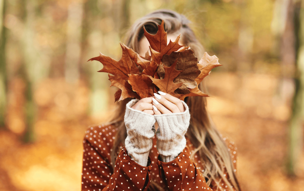 Woman in a woods setting is holding some fallen maple leaves in front of her face.