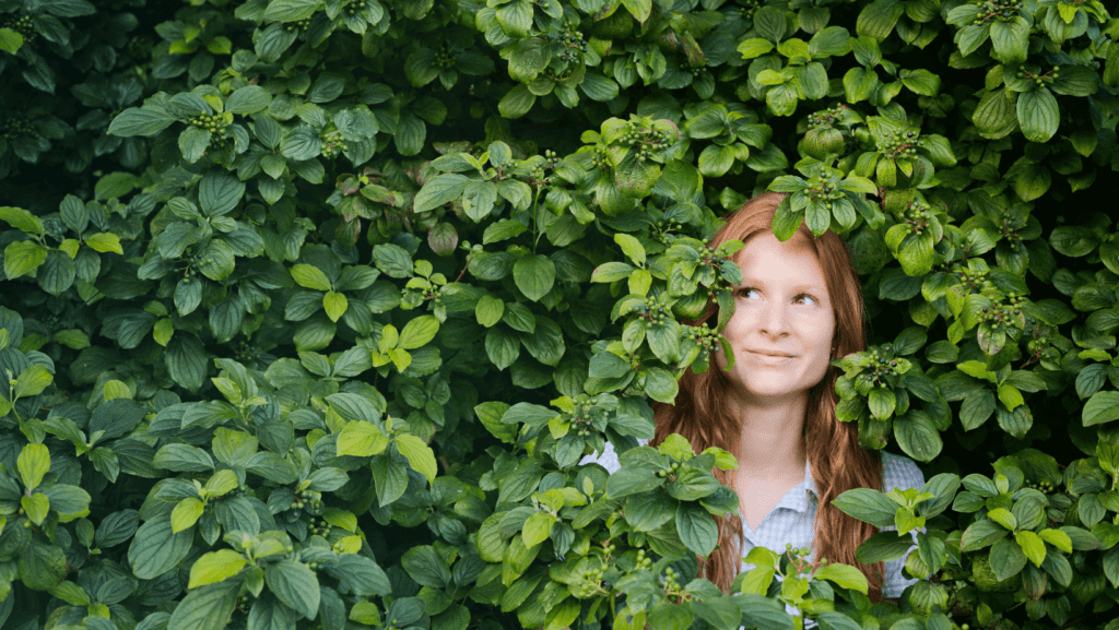 A woman with beautiful red hair is looking up while standing in green hedges almost covering her completely.